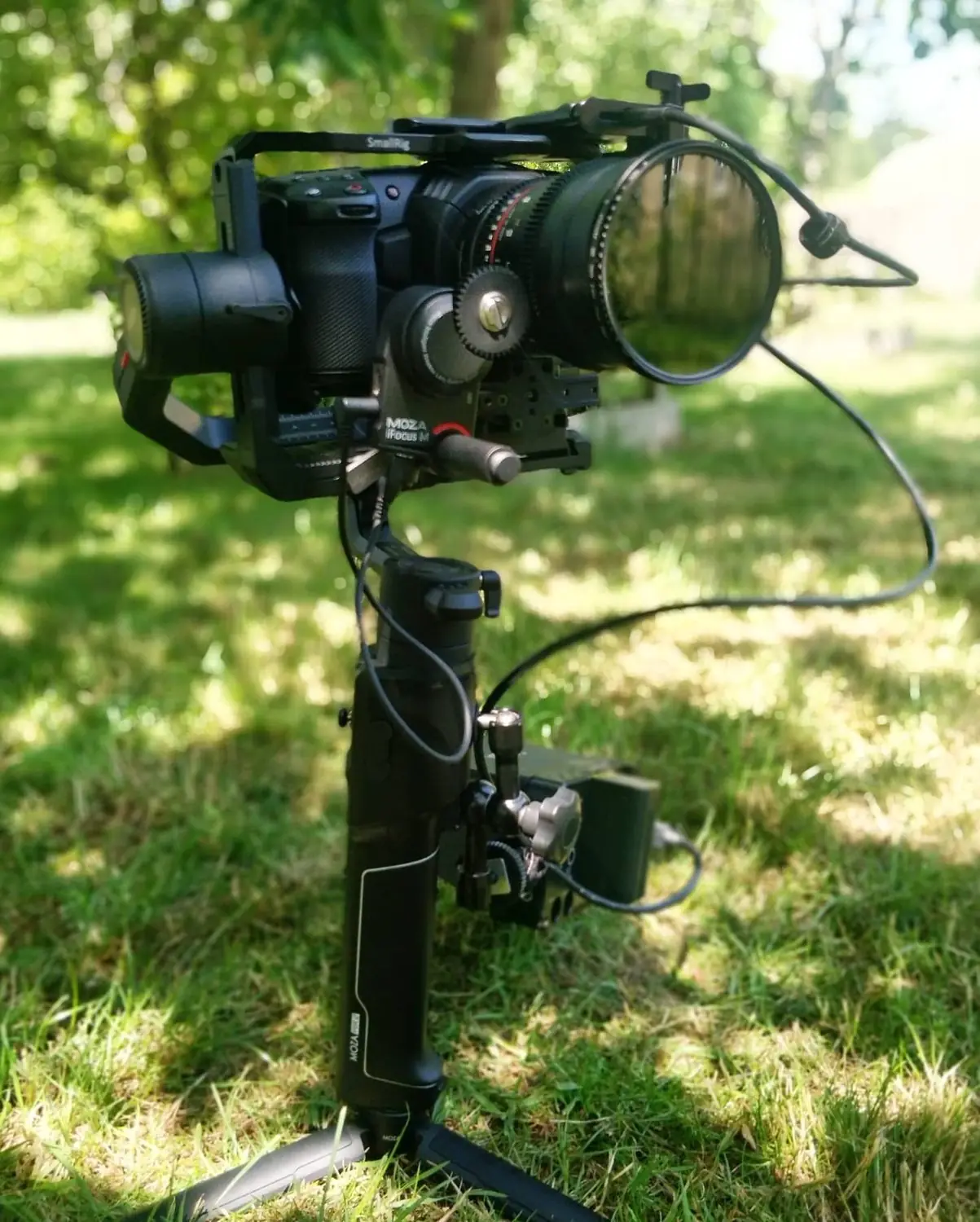 A camera on a gimbal stabiliser standing on a tripod in a sunlit field, with trees behind