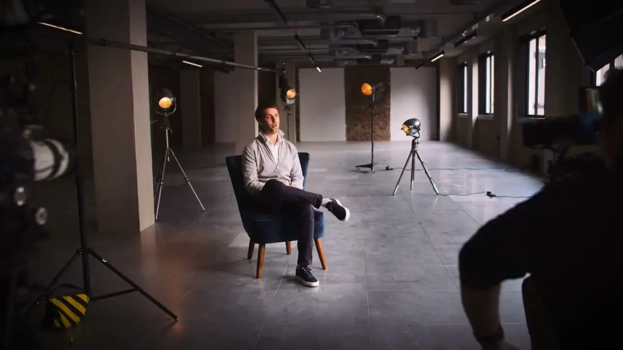 A man in an armchair being interviewed in a large bare studio, lamps on stands and a camera in the foreground