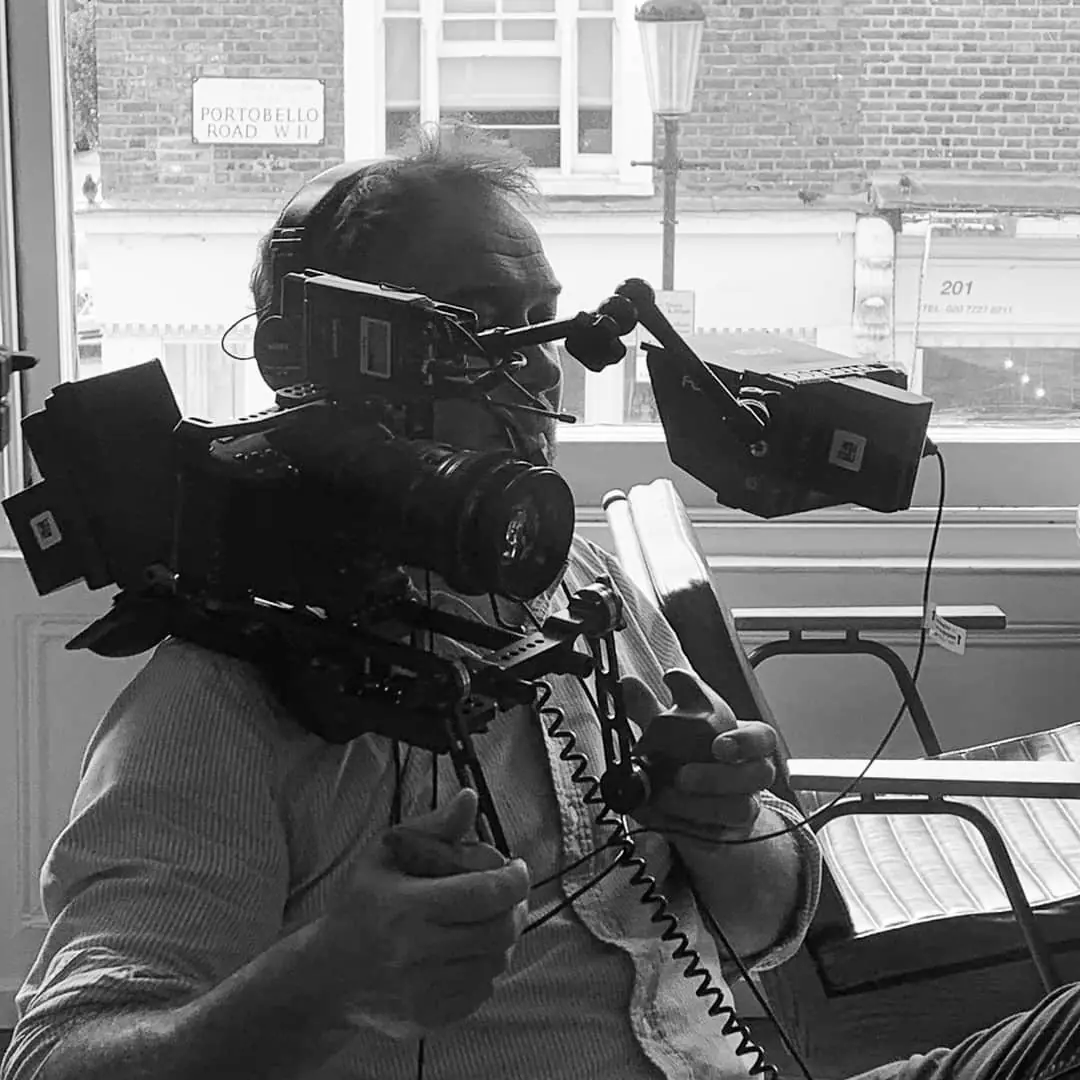 A black and white photograph of an operator behind a shoulder-mounted camera, working beside a window