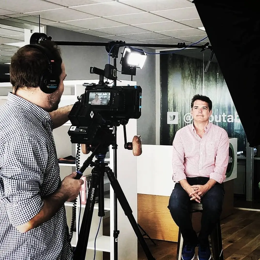 A crew filming an interview: a camera operator with headphones at a tripod and a man in a pink shirt sitting on a stool