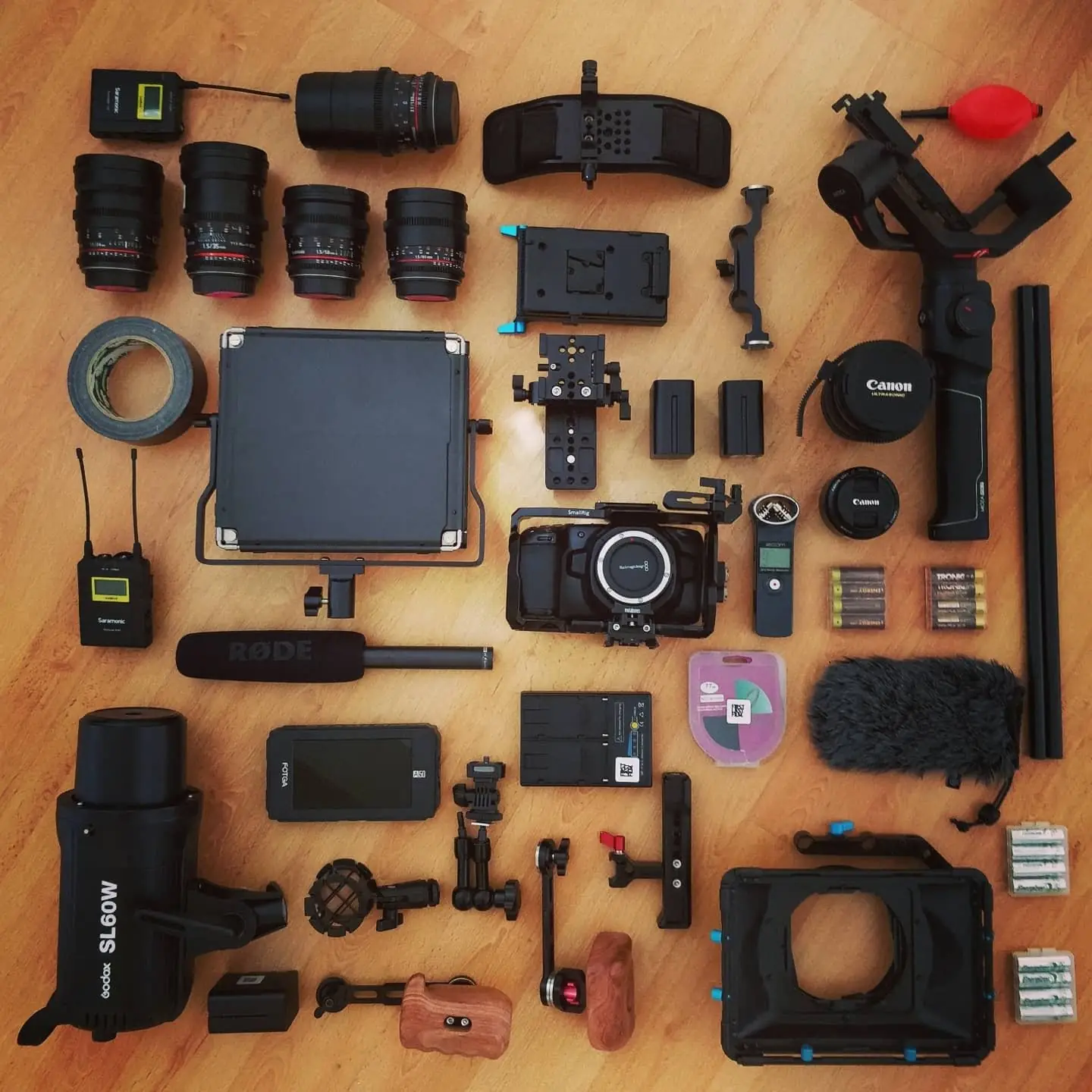 The whole kit laid out in a grid on a wooden floor: lenses, a gimbal, a matte box, radio mics, batteries, a monitor and cables