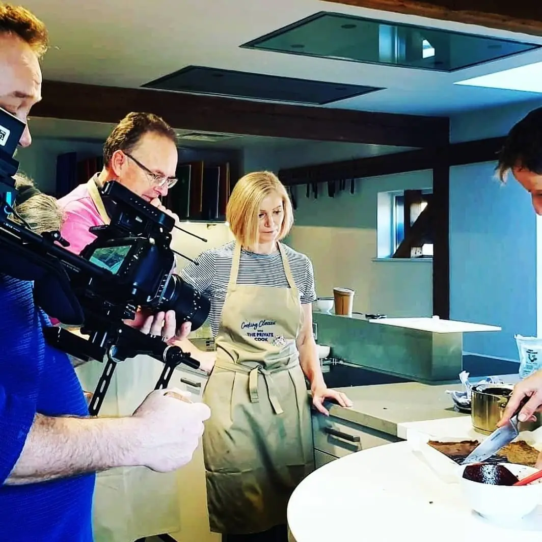 A crew filming in a kitchen: a camera operator and a boom operator with a woman in an apron at a worktop