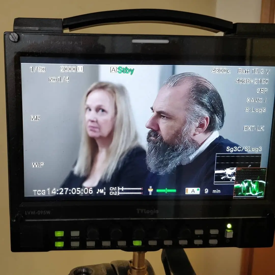A field monitor on set showing a two-shot of a woman and a bearded man in an interview, with scopes in the corner