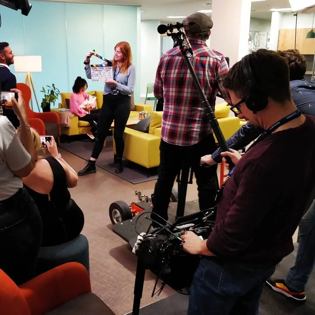 A crew in a bright office filming a scene with a clapperboard, a slider and a camera operator in the foreground