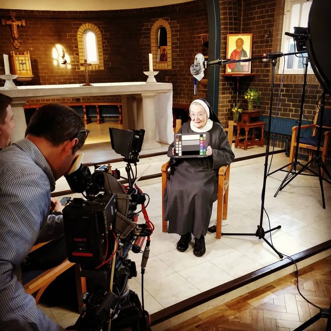 A filming set in a small chapel, with a camera and lights pointed at an elderly nun seated on a chair
