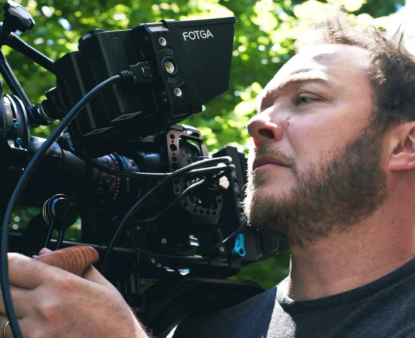A bearded camera operator looking intently through a camera rig fitted with a monitor, trees behind him
