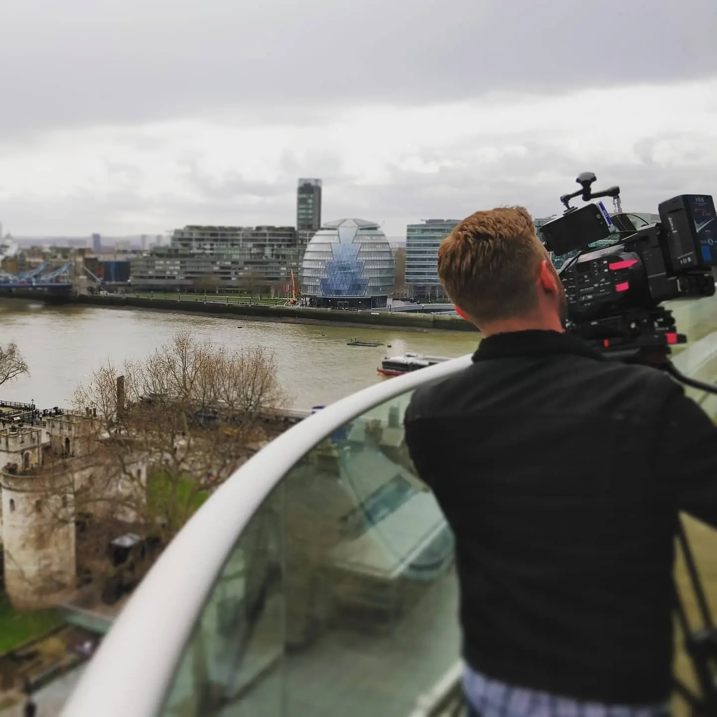 A camera operator in a dark jacket filming from a glass balcony over the Thames, with the city on the far bank