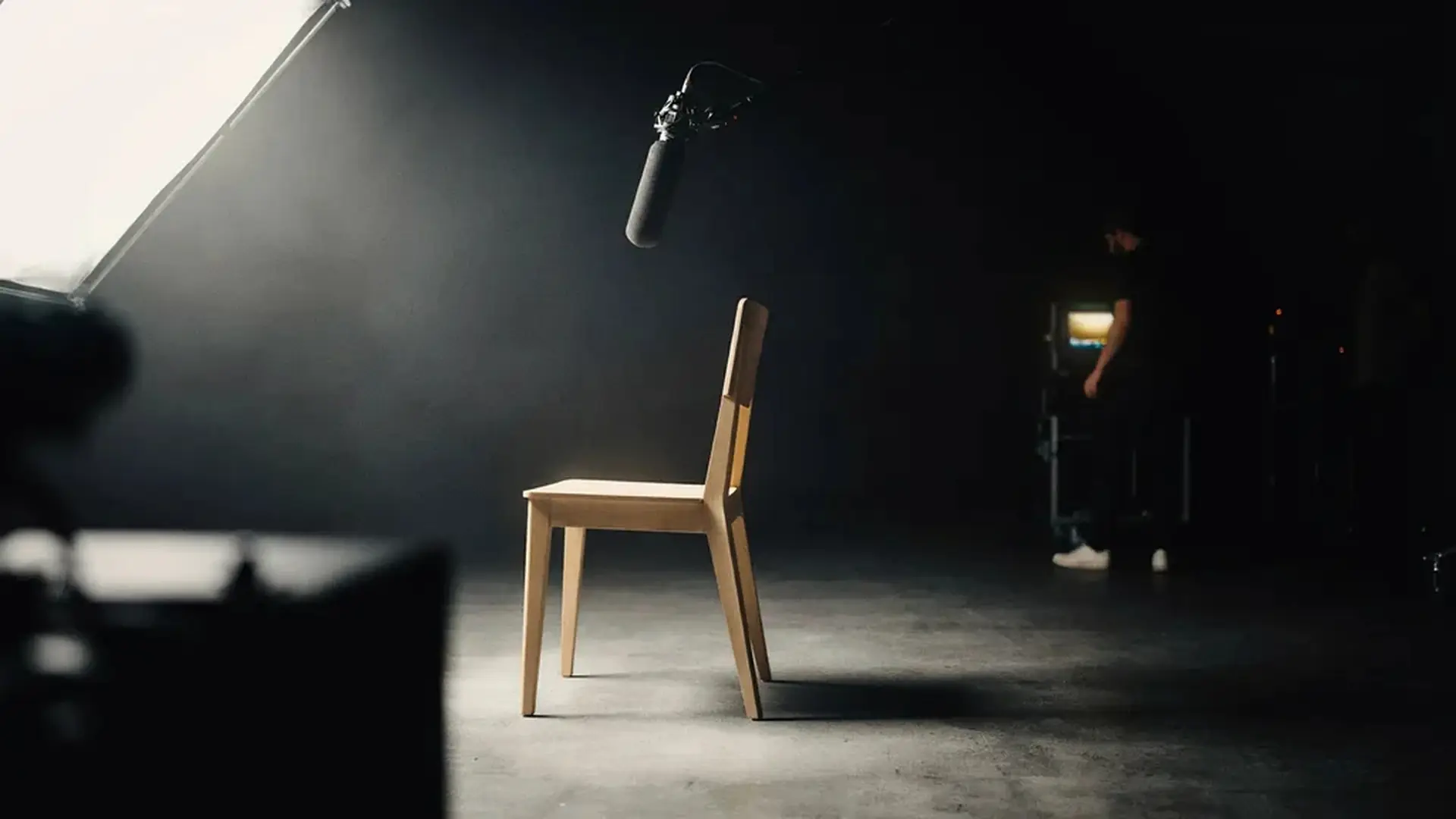 An empty wooden chair lit by a single shaft of light in a dark studio, with a boom microphone overhead and a crew member in the background