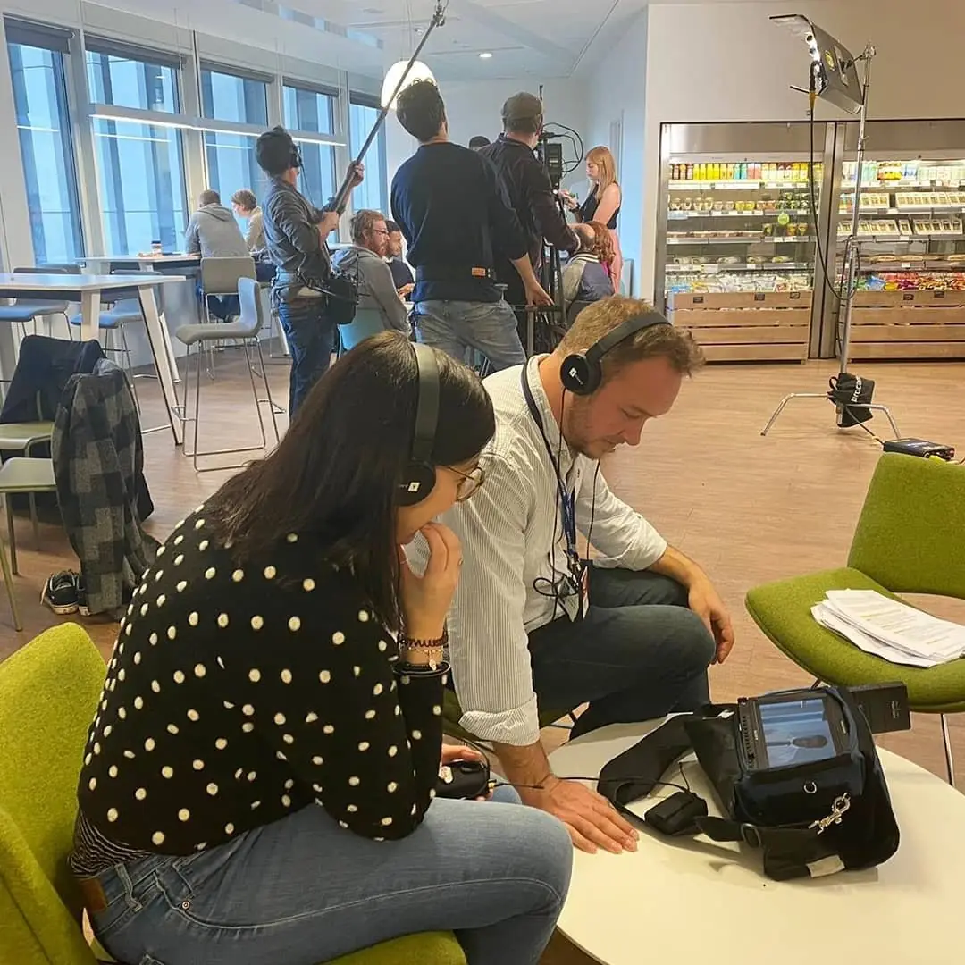 A sound recordist with headphones and a woman in a polka dot top at a monitor on a shoot in a bright shop interior