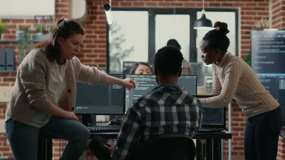 Two women leaning in to point at code on a bank of monitors in a brick-walled office, a seated man between them