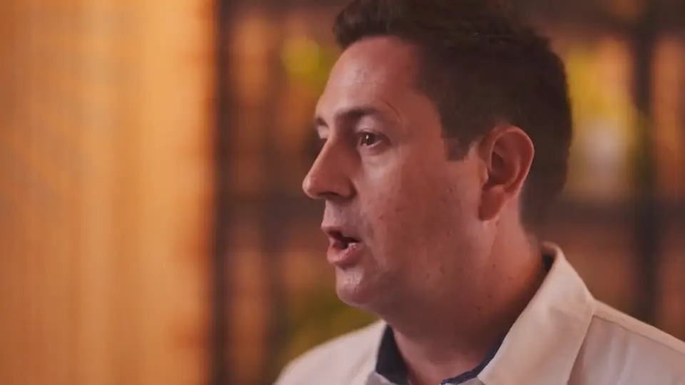Close side view of a man in a white shirt speaking, with a warm blurred restaurant interior behind him