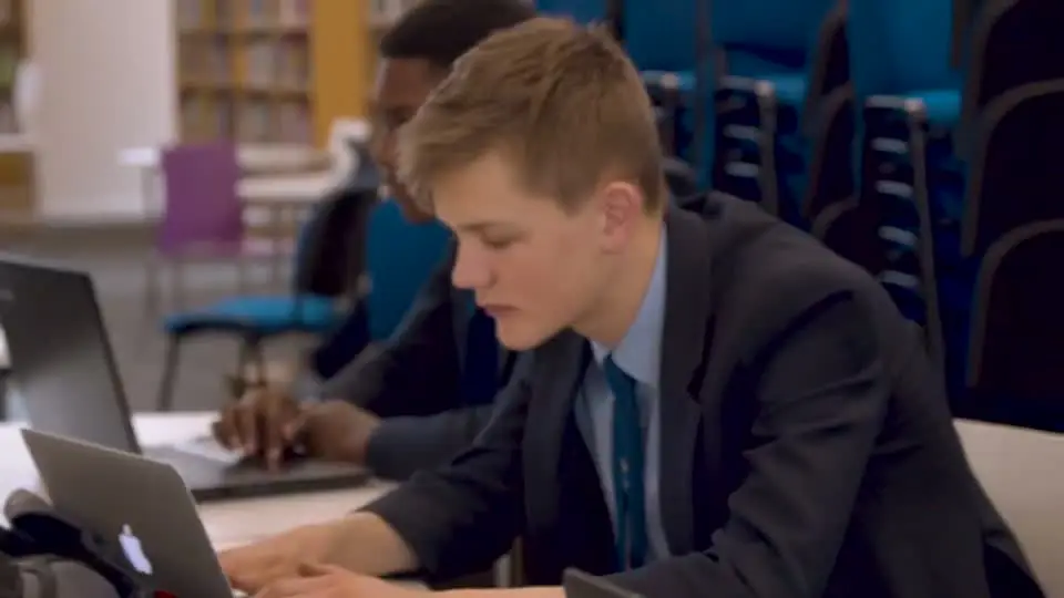 A schoolboy in a blazer typing on a laptop at a library desk, other pupils beside him