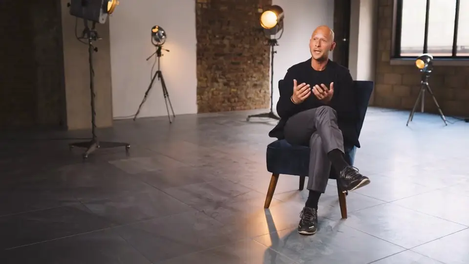 A bald man in black sitting on a navy armchair in a bare studio, gesturing as he talks, with tungsten lamps on stands behind