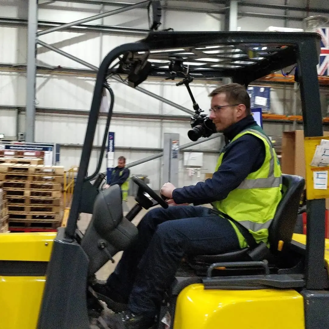 A man in a hi-vis vest riding a forklift in a warehouse, with a camera mounted on the forklift roof
